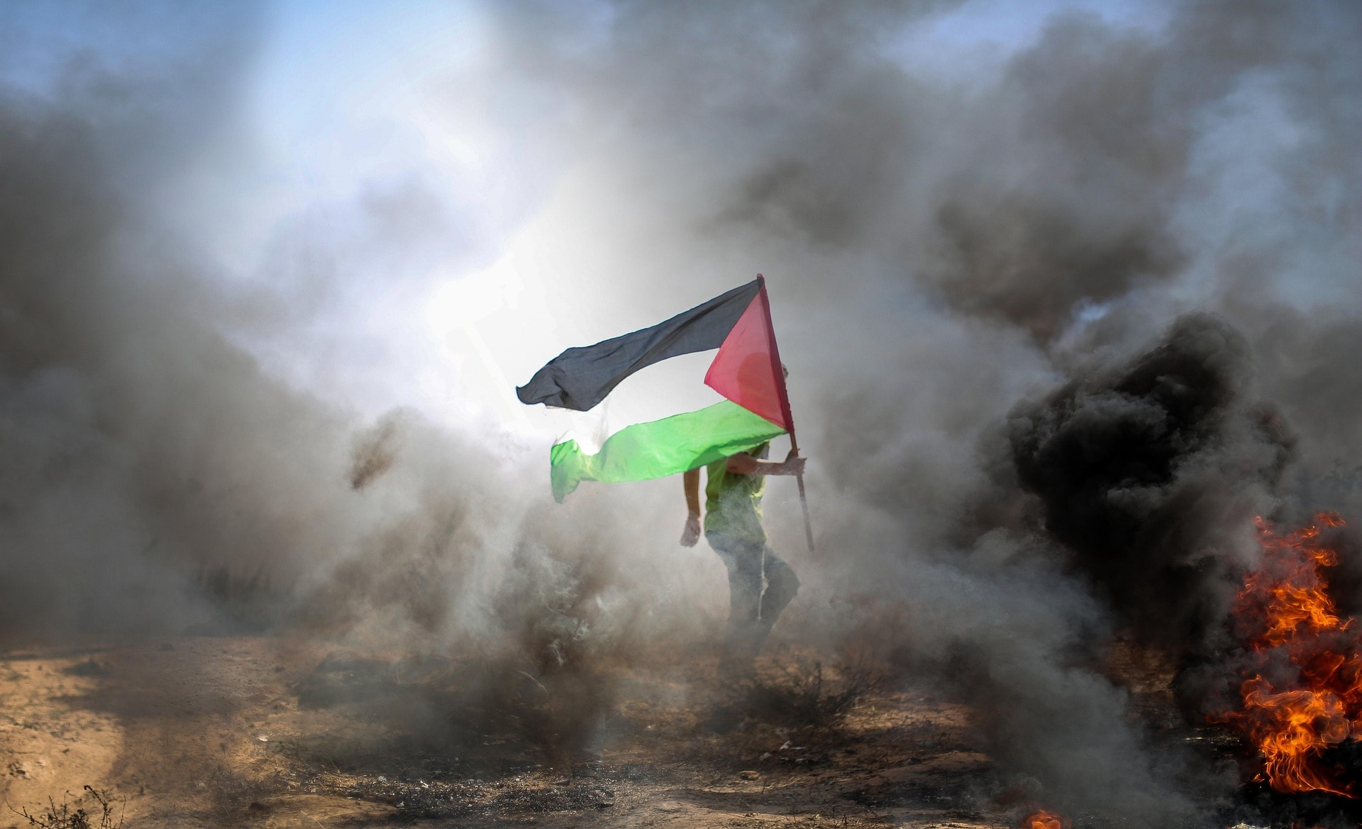 Em meio à fumaça e às chamas, homem empunha bandeira da Palestina. (Imagem ilustrati va – fonte: Pxfuel, domínio público)