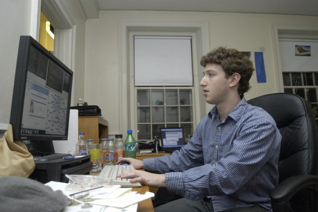 Mark Zuckerberg em seu dormitório em Harvard, em 2004, após lançar o TheFacebook.com. O retrato do “jovem gênio” consolidou a narrativa de sucesso individual que legitima a desigualdade e a concentração de poder das Big Techs. Crédito: Lowell K. Chow. Imagem de uso editorial sob direitos de The Harvard Crimson.