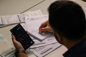 Pessoa calcula despesas domésticas diante de contas e boletos, em imagem que retrata os desafios do orçamento familiar e do endividamento no Brasil. Foto: Joédson Alves/Agência Brasil.